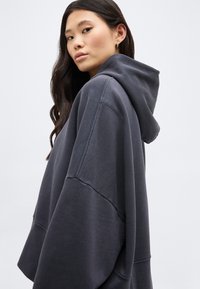 Νεαρή γυναίκα με μακριά σκούρα μαλλιά, φοράει υπερμεγέθη σκουρόχρωμη γκρι φούτερ, κοιτάζοντας πλάγια, μπροστά σε ένα απλό φωτεινό φόντο.