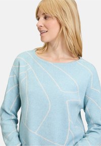Lichtblauwe trui van zacht materiaal, met een geometrisch wit patroon, ronde halslijn en lange mouwen.