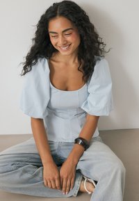 Himmelblaue Bluse mit Puffärmeln, quadratischem Ausschnitt und subtilen vertikalen Nähten, kombiniert mit hochtaillierten hellen Jeans und einem dunklen Armband.