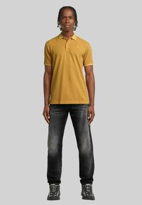 Polo di colore giallo senape con colletto e maniche corte, abbinato a jeans grigio scuro e sneaker nere. Design semplice e casual.