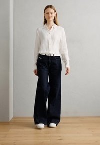 Chemise blanche à manches longues et boutons, jeans larges sombres et baskets blanches. Le tissu lisse et la coupe droite mettent en valeur les lignes épurées de l'ensemble.