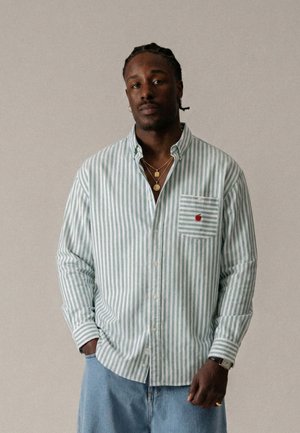 Mężczyzna z warkoczami, ubrany w zielono-białą koszulę w paski na guziki z logo Apple na kieszeni, jasnoniebieskie dżinsy i złote łańcuszki.