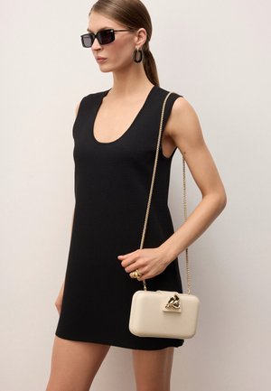 Žena v černých bezrukávových šatech a velkých černých náušnicích nese malou krémovou kabelku se zlatým řetízkem a zapínáním, má na sobě černé sluneční brýle.