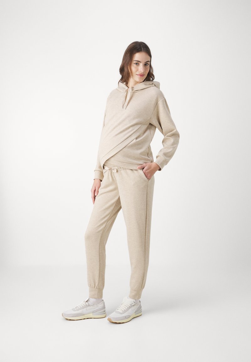 Beige huvtröja med crossover-design, parat med matchande joggers. Materialet verkar mjukt och bekvämt, med elastiska manschetter.