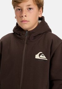 Hnědá mikina na zip s vysokým límcem, fleecovou podšívkou a krémovým logem Quiksilver. Má hladký povrch a standardní střih.