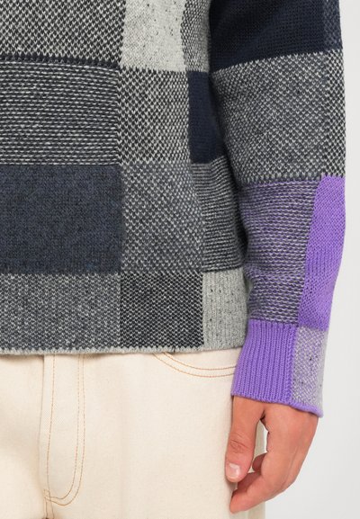 Suéter de punto patchwork en tonos de gris y púrpura en una persona que lleva pantalones beige claros, con un enfoque en la manga y la mano.