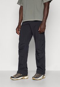 Mustat kevyet cargo-housut vetoketjuilla, joissa on sileä pinta ja toiminnallisia taskuja, yhdistettynä tummiin, karkeisiin urheilukenkäisiin.