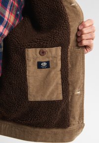 Giacca in velluto a coste marrone con fodera in pelliccia marrone morbida, dotata di una tasca beige con un bottone di legno e una etichetta del marchio all'interno.
