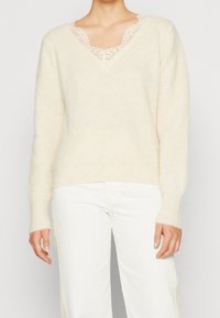 Crème gebreide trui met V-hals, benadrukt door kanten rand. Lange mouwen, zachte textuur, gecombineerd met witte broek.