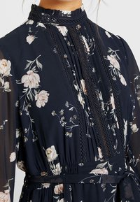 Nära bild av en modell som bär en mörk blommig klänning med intrikata spettdetaljer och veck, med hög hals och mjuk, flödande tyg.