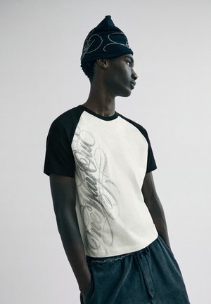 Giovane uomo con maglietta raglan bianca e nera con testo in corsivo, berretto blu navy con disegni e pantaloni larghi scuri, che posa davanti a uno sfondo plain.