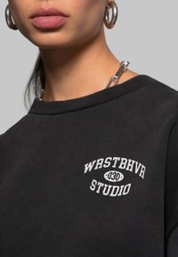 Schwarzes Sweatshirt mit geripptem Rundhalsausschnitt, versehen mit dem weißen Textlogo "WRSTBHVR 030 STUDIO". Glatte Textur und lässiger Schnitt ohne sichtbare Muster.