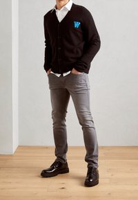 Man som bär en svart cardigan med en blå "W"-märke, vit skjorta, grå smala jeans och svarta snörskor, står på ett trägolv.
