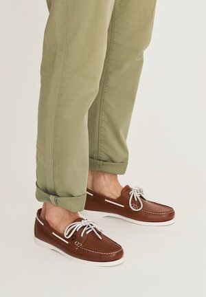 Man draagt opgerolde olijfgroene broek en bruine leren bootschoenen met witte veters op een lichte achtergrond.