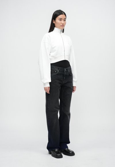 Felpa bianca cropped con zip, jeans neri a vita alta e scarpe nere. Modella in piedi su uno sfondo chiaro e neutro.