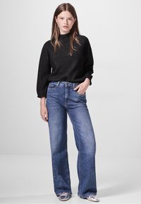 Maglione nero a maniche lunghe con finitura testurizzata e collo alto, abbinato a jeans larghi azzurri e scarpe argentate metalliche.
