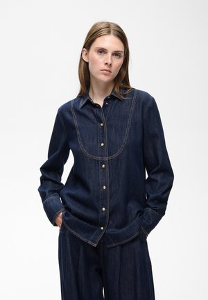 Femme debout les mains dans les poches, portant une chemise en jean bleu foncé et un pantalon large assorti sur un fond blanc uni.