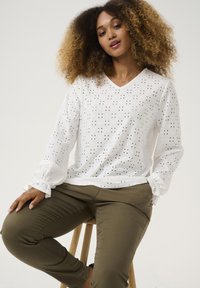 Blusa blanca de manga larga con patrón de eyelet abierto, escote en V, puños fruncidos, combinada con pantalones verde oliva, sentada en un taburete de madera.