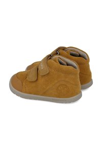 Botas de ante marrón hasta el tobillo con punta redonda, dos correas de gancho y bucle, parte superior texturizada y suelas de goma grises; presenta un logo en relieve.