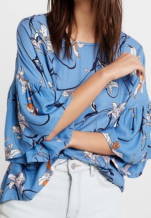 Femme portant un chemisier bleu clair à motifs floraux avec manches 3/4 volantées et un pantalon blanc taille haute, bras croisés à la taille.