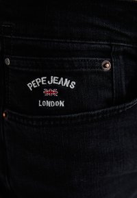 Mustad denim teksad, millel on seljatasku, kus on valge tikand "PEPE JEANS" ja "LONDON", mida kaunistab väike Ühendkuningriigi lipp.