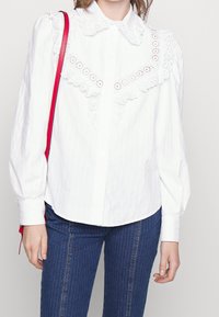 Camicia bianca con colletto in pizzo a scallop e dettagli ricamati intricati. Abbinata a pantaloni in denim a righe blu.