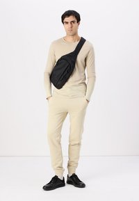 Svart slingbag brukt over kroppen over en lys beige langermet skjorte og matchende joggebukser; med glidelåser og en teksturert overflate.