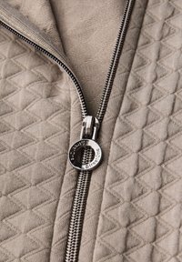 Ljust beige kviltad tyg med ett diamantmönster, med en silverfärgad dragked jag med en cirkulär ring och varumärkesdetaljer.