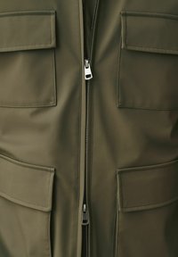 Olivengrøn jakke foran med to øvre og to nedre klaplommer samt en central sølvfarvet dobbeltlynlås delvist lynet op.