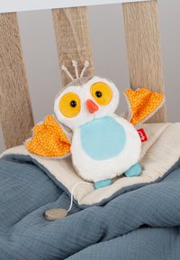 Jouet hibou en peluche avec de grands yeux jaunes, des ailes orange à pois et un ventre bleu reposant sur des couvertures bleues et beiges superposées sur une chaise en bois.
