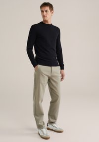 Hombre de pie con las manos en los bolsillos, vistiendo un suéter negro, pantalones beiges y zapatillas blancas sobre un fondo claro.