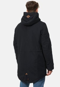 Marinblå långrock med huva, i ett texturerat tyg, med bruna läderdetaljer och en triangulär detalj vid fållen, fäst med en knapp.