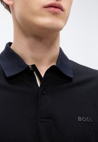 Μαύρο πόλο μπλουζάκι με ναυτικό γιακά, δύο κουμπιά και υφασμάτινη υφή. Διαθέτει διακριτικό ριγέ μοτίβο και ανάγλυφο λογότυπο στο στήθος.