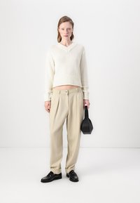Elfenbensfarget strikkegenser med V-hals, paret med beige plisserte bukser. Holder en svart rektangulær veske og har på seg svarte sko.