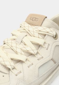 Μπεζ σουέντ αθλητικό παπούτσι με λευκά κορδόνια με σχέδιο, μικρό λογότυπο UGG, στοιχεία από δίχτυ και λεπτομέρειες αντίθετης ραφής.