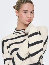 Krämfärgad ribbstickad tröja med svarta horisontella ränder, hög krage och långa ärmar, bärs med stora silverörhängen i ring.