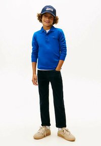 Blauw poloshirt met lange mouwen, donkerblauwe broek, beige sneakers en een donkerblauwe pet met witte letters, staand tegen een effen achtergrond.
