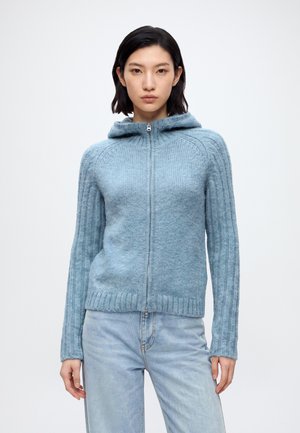 Femme portant un sweat à capuche zippé en maille bleu clair avec des manches côtelées et un jean bleu clair, debout devant un fond blanc uni.