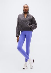 Γκρι φλις ζακέτα με φερμουάρ και μπροστινή τσέπη, συνδυασμένη με εφαρμοστά μωβ-ανοιχτό κολάν και λευκά αθλητικά παπούτσια με σκούρες λεπτομέρειες.