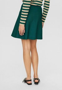 Vihreä liehuva hame, joka on valmistettu sileästä materiaalista ja jossa on hyvin istuva vyötärö. Yhdistetty tyylikkäisiin mustiin sandaaleihin, joissa on yksinkertainen muotoilu.