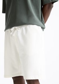 Shorts en coton blanc avec une taille élastique, un cordon de serrage et des poches latérales. Associés à un t-shirt vert foncé à manches courtes.