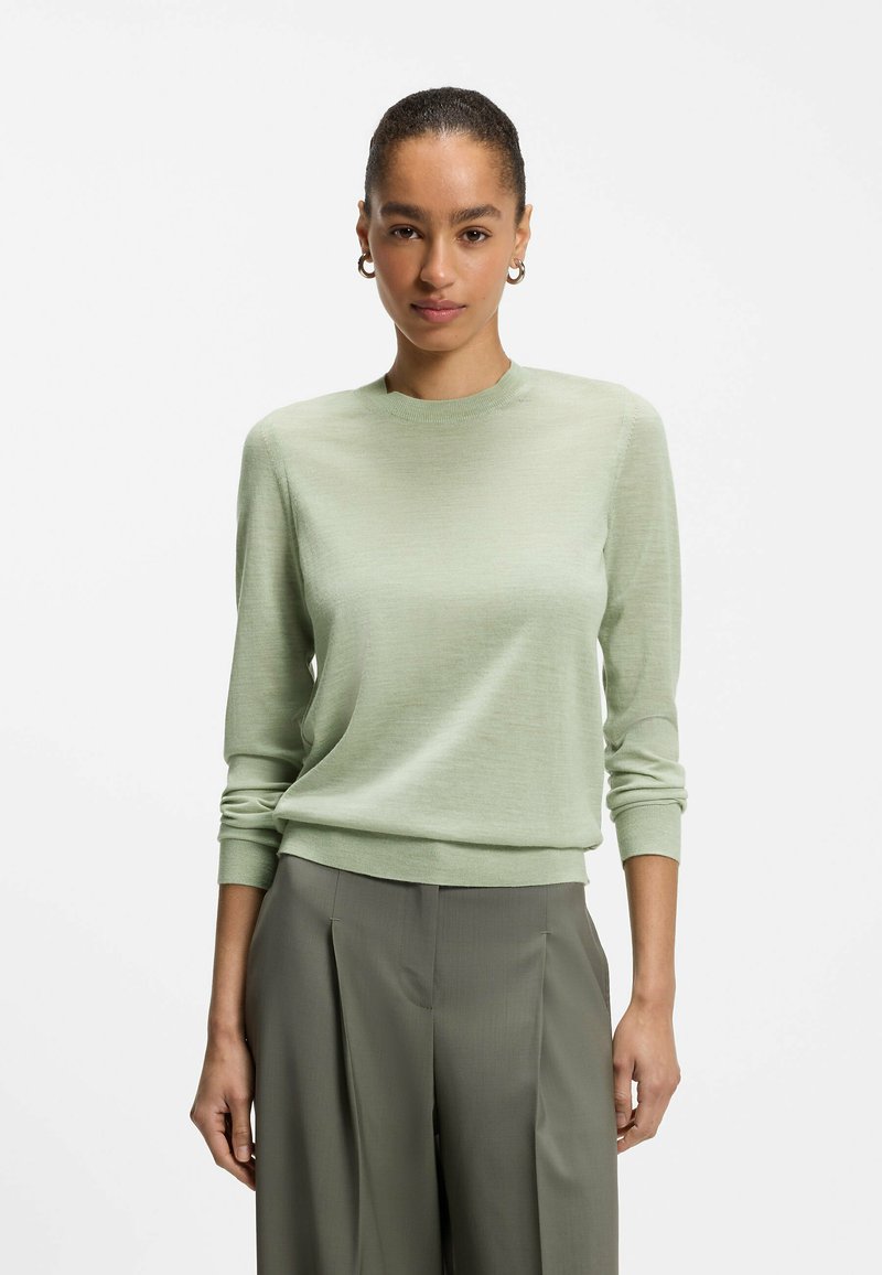 Jeune femme aux cheveux tirés en arrière porte un pull vert clair et un pantalon plissé vert foncé taille haute, debout devant un fond uni.