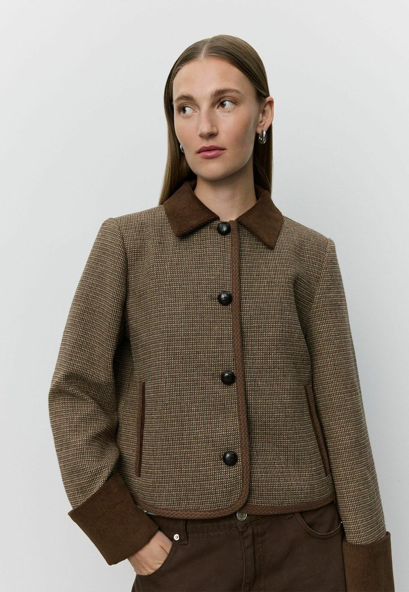 Femme aux cheveux raides porte une veste marron à carreaux avec un col, des poignets et des boutons marron foncé, une main dans la poche, sur un fond uni.