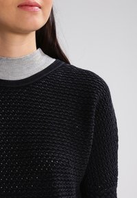 Eine Frau trägt einen schwarzen strukturierten Strickpullover über einem hellgrauen gerippten Rollkragenpullover, sichtbar von Nacken bis Schulter vor einem grauen Hintergrund.