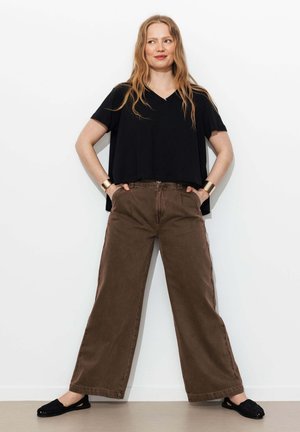Femme debout contre un mur blanc, portant un haut noir à manches courtes, un pantalon large marron, des chaussures noires et des bracelets dorés, les mains dans les poches.