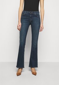 Vaqueros acampanados de color azul oscuro en tela de denim, con una cintura de altura media y bolsillos frontales. Combinados con tacones de gamuza marrón. Diseño sencillo y clásico.