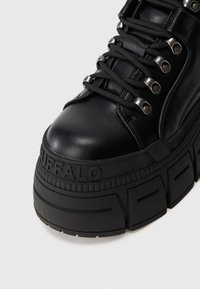 Melnas augstās kedas ar masīvu zoli, gludu ādas virsu, metāla acīm un bieziem melnajiem šņorēm. Zīmols redzams sānos.