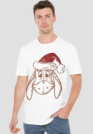 Man wearing white t-shirt with a smiling cartoon dog face wearing a red Santa hat, standing with one hand in pocket.