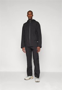 Veste noire imperméable avec fermeture éclair frontale et capuche ajustable, associée à un pantalon noir assorti et des baskets de couleur claire.