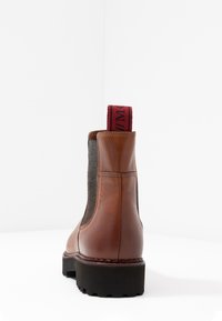 Bottine en cuir marron avec des panneaux latéraux en tissu gris, semelle texturée et tirant rouge. Dotée d'un bout rond et de coutures contrastées.